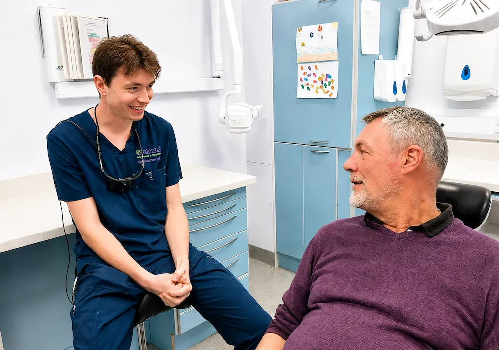 Young male dentist in blue scrubs talking and smiling with older male patient in dental chair.