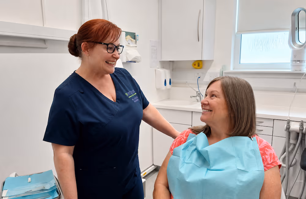 Smiling female dental professional talks with female patient in dental clinic room with white cabinets.