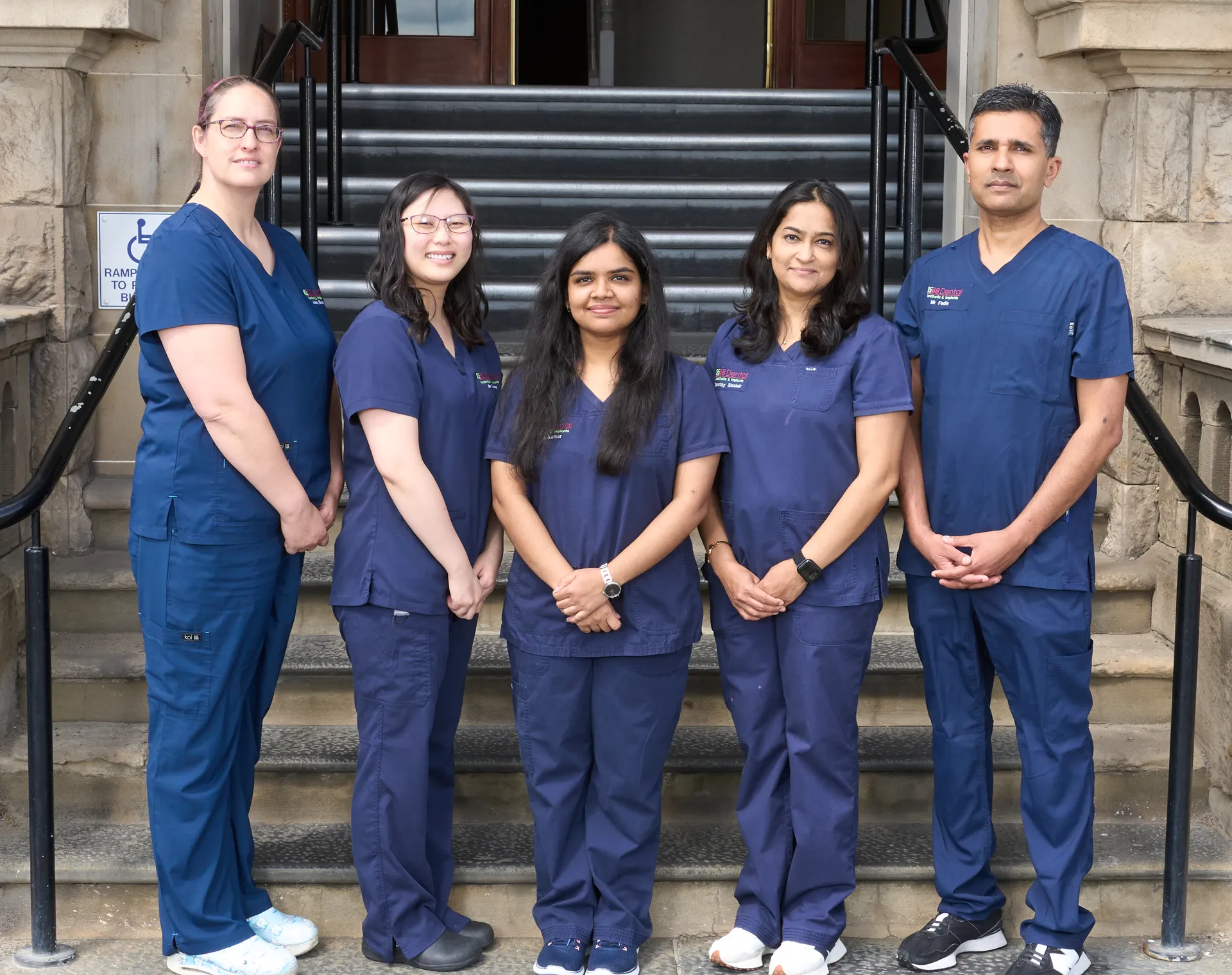 Five healthcare professionals in navy scrubs standing on steps outside a building, smiling.