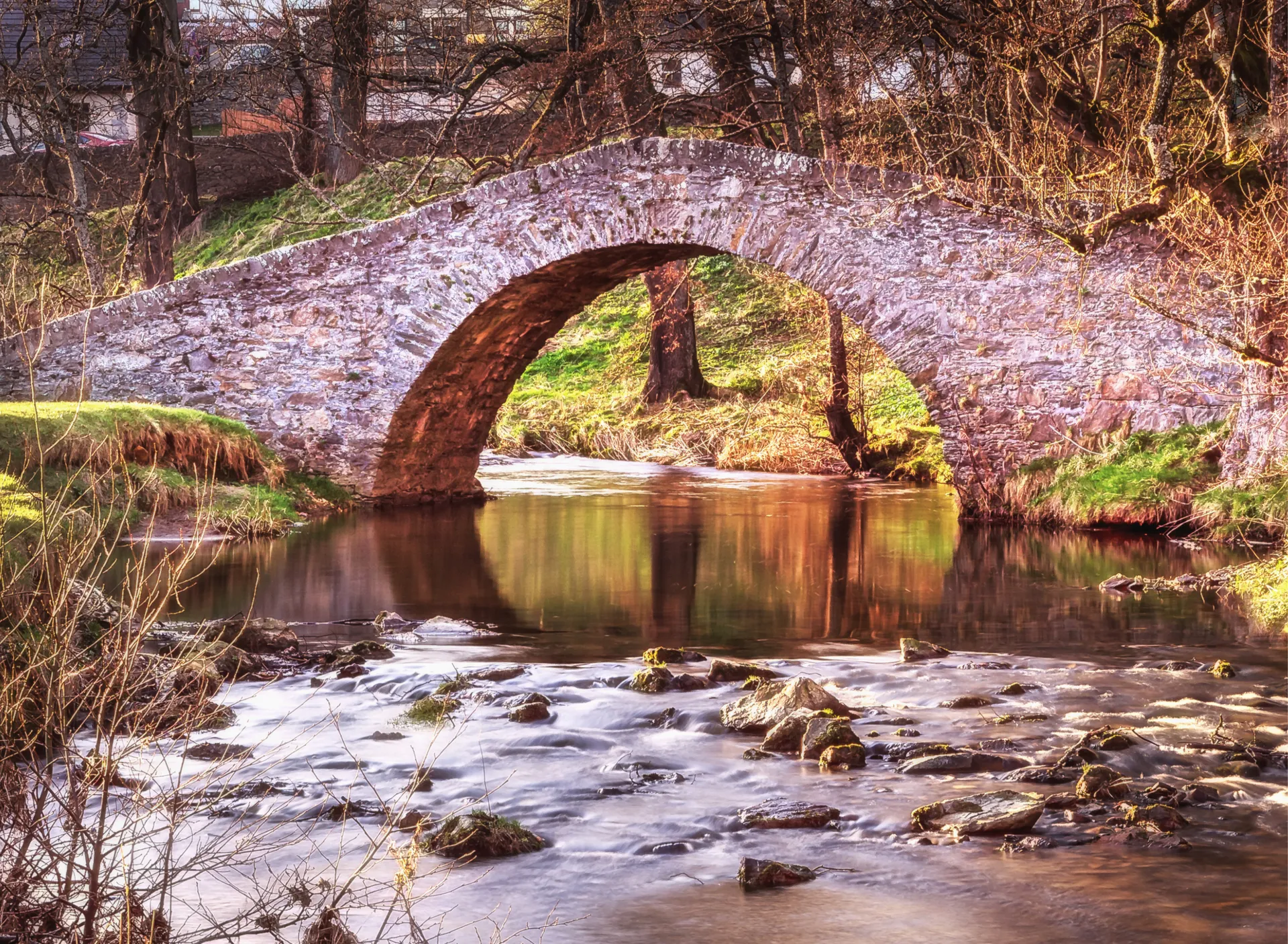 Old stone arch bridge over a calm river with rocks and trees in a sunlit rural setting.