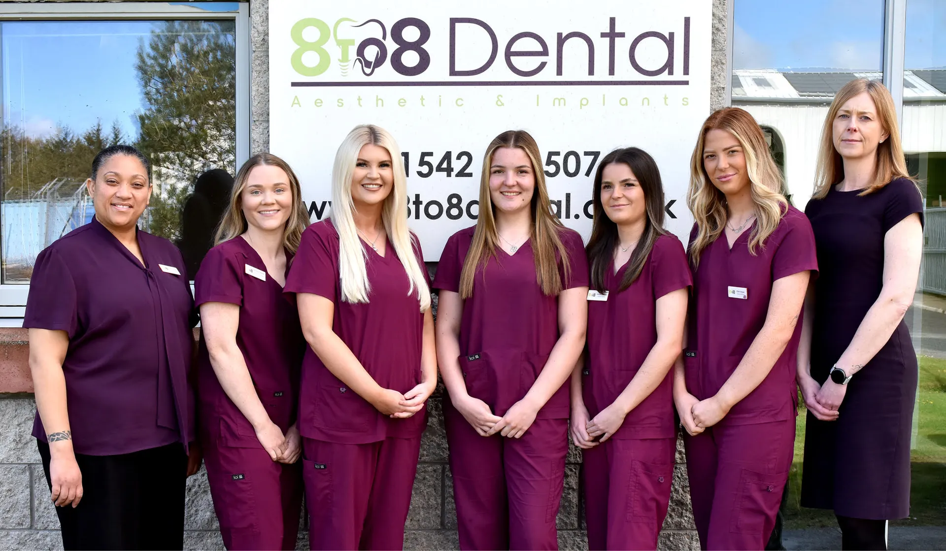 Seven female dental staff standing in front of a dental clinic sign, wearing purple uniforms.
