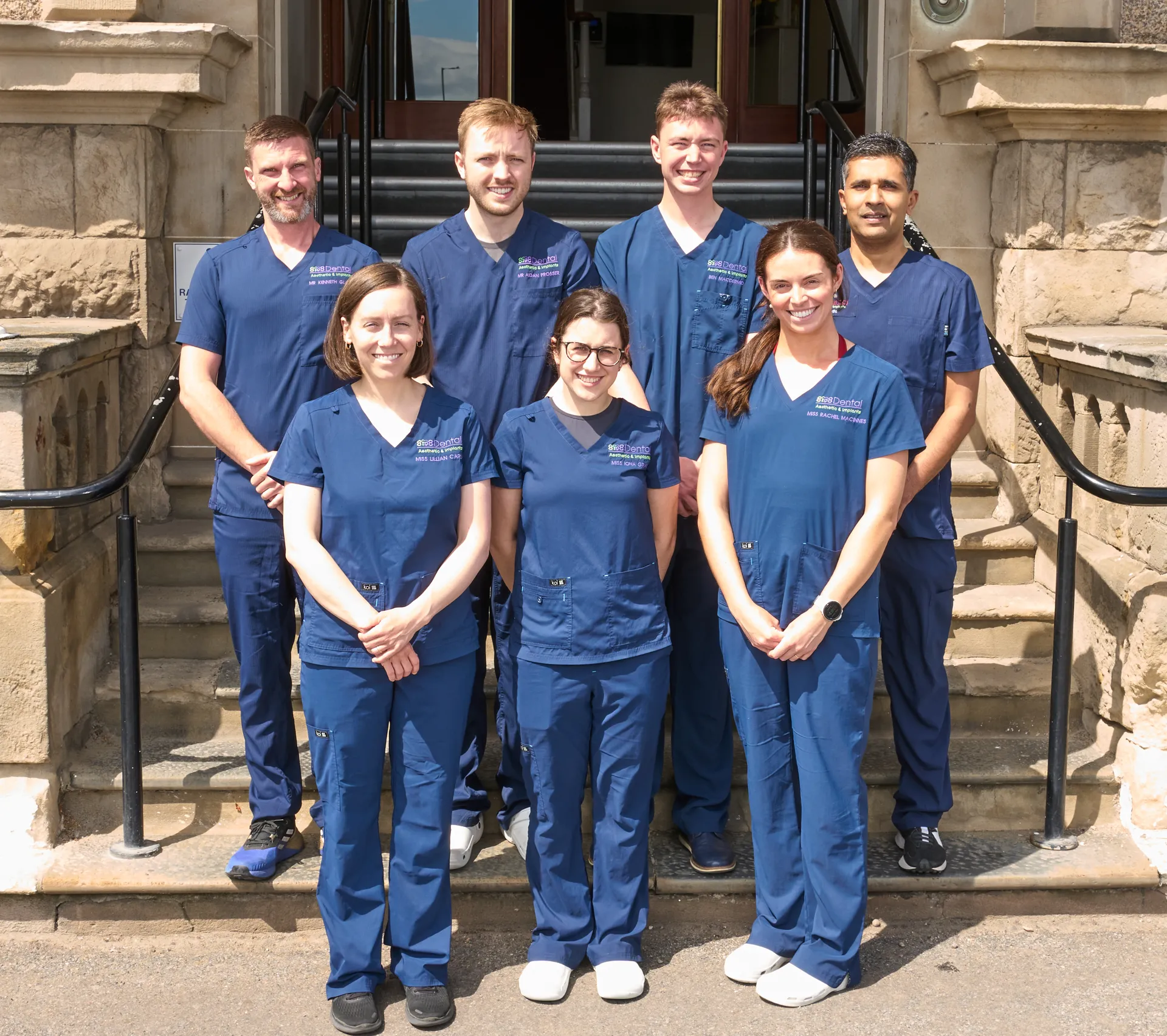 Seven dental professionals wearing blue scrubs posing on outdoor stone steps in front of a building.