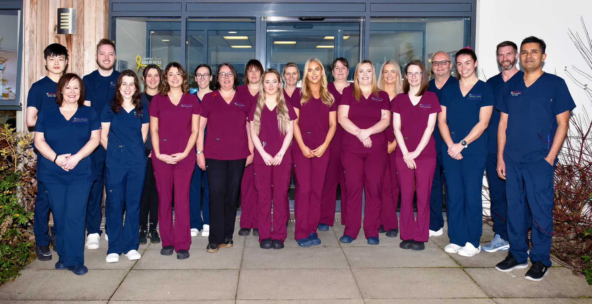 Group of medical staff in maroon and navy scrubs standing outside a building entrance.