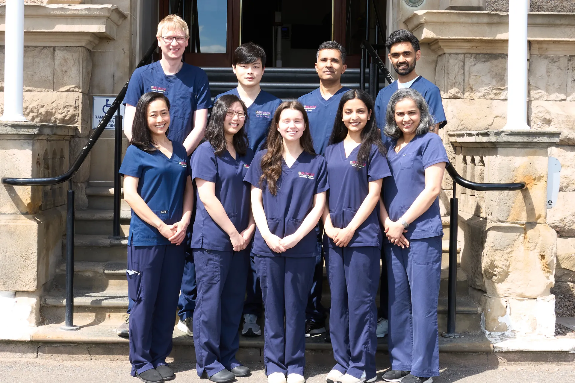 Group of nine diverse medical staff in blue scrubs standing on stone steps outside building.