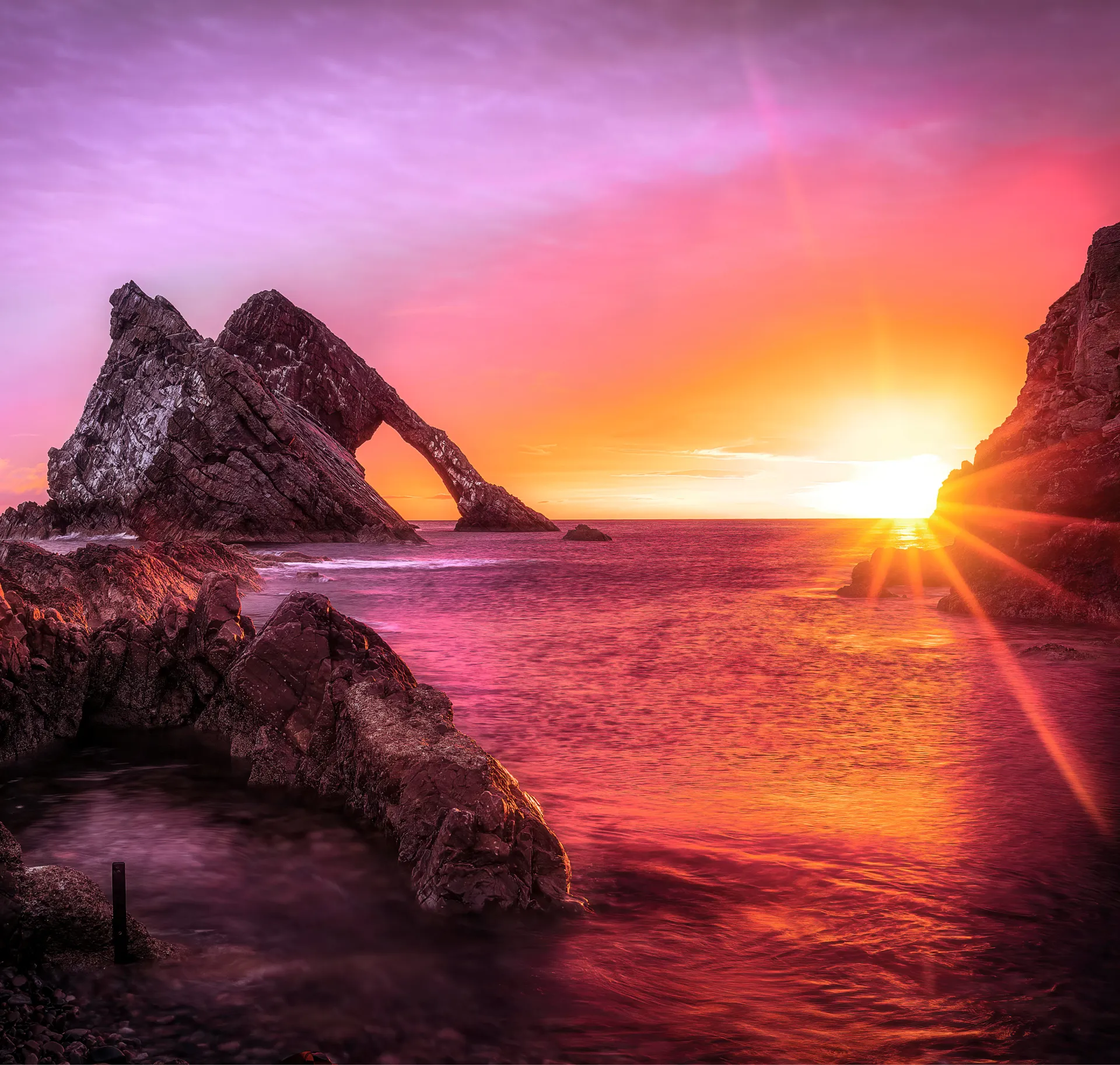 Sunset over ocean with rocky formations, including a large arch-shaped rock on the left.
