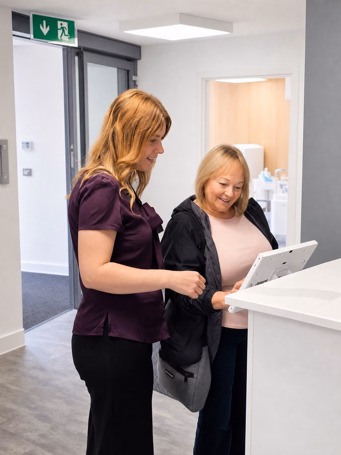 Two women smiling and looking at a screen on a white desk in a modern office.