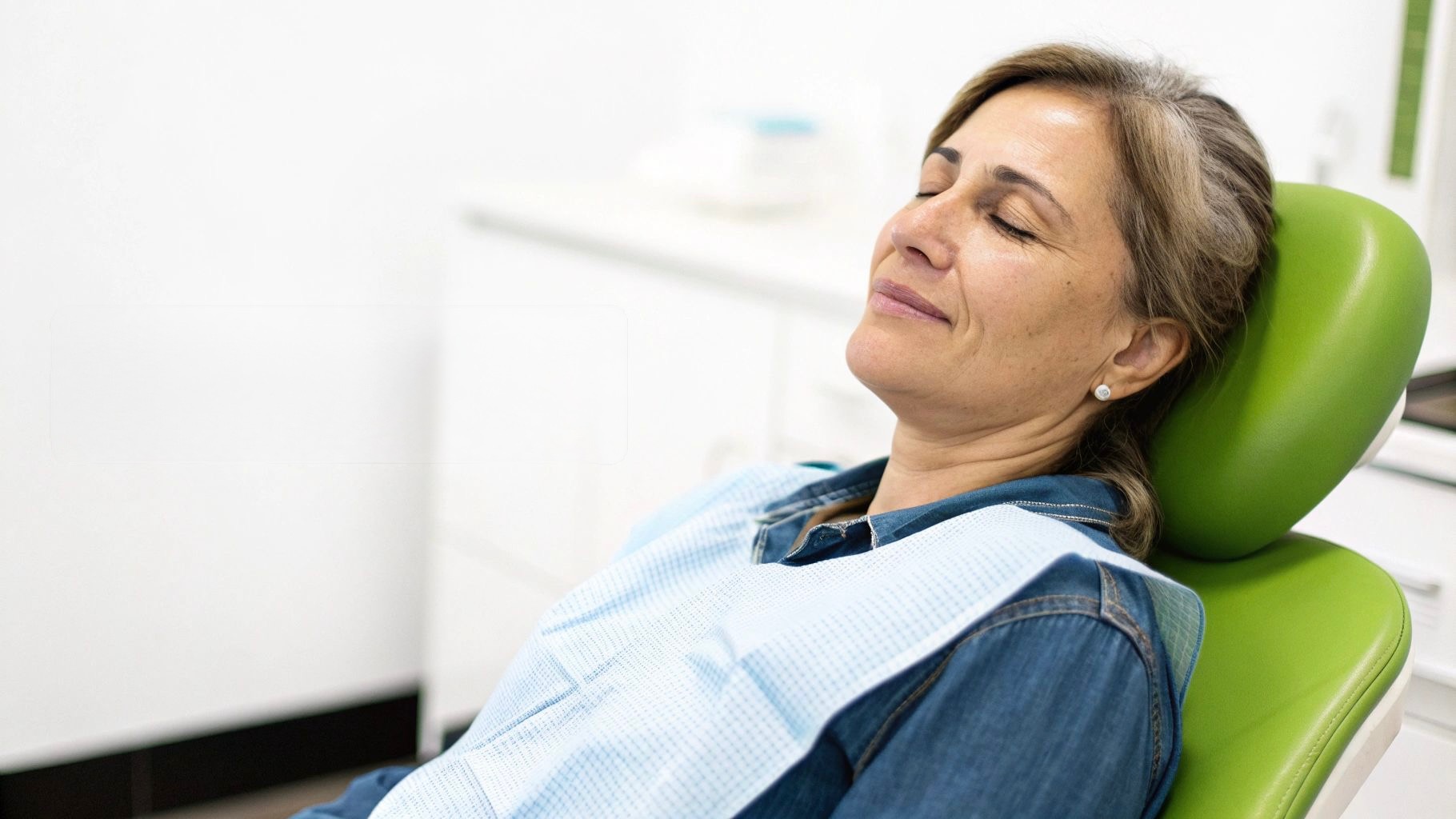 Relaxed woman with eyes closed sitting in a green dental chair wearing a dental bib.