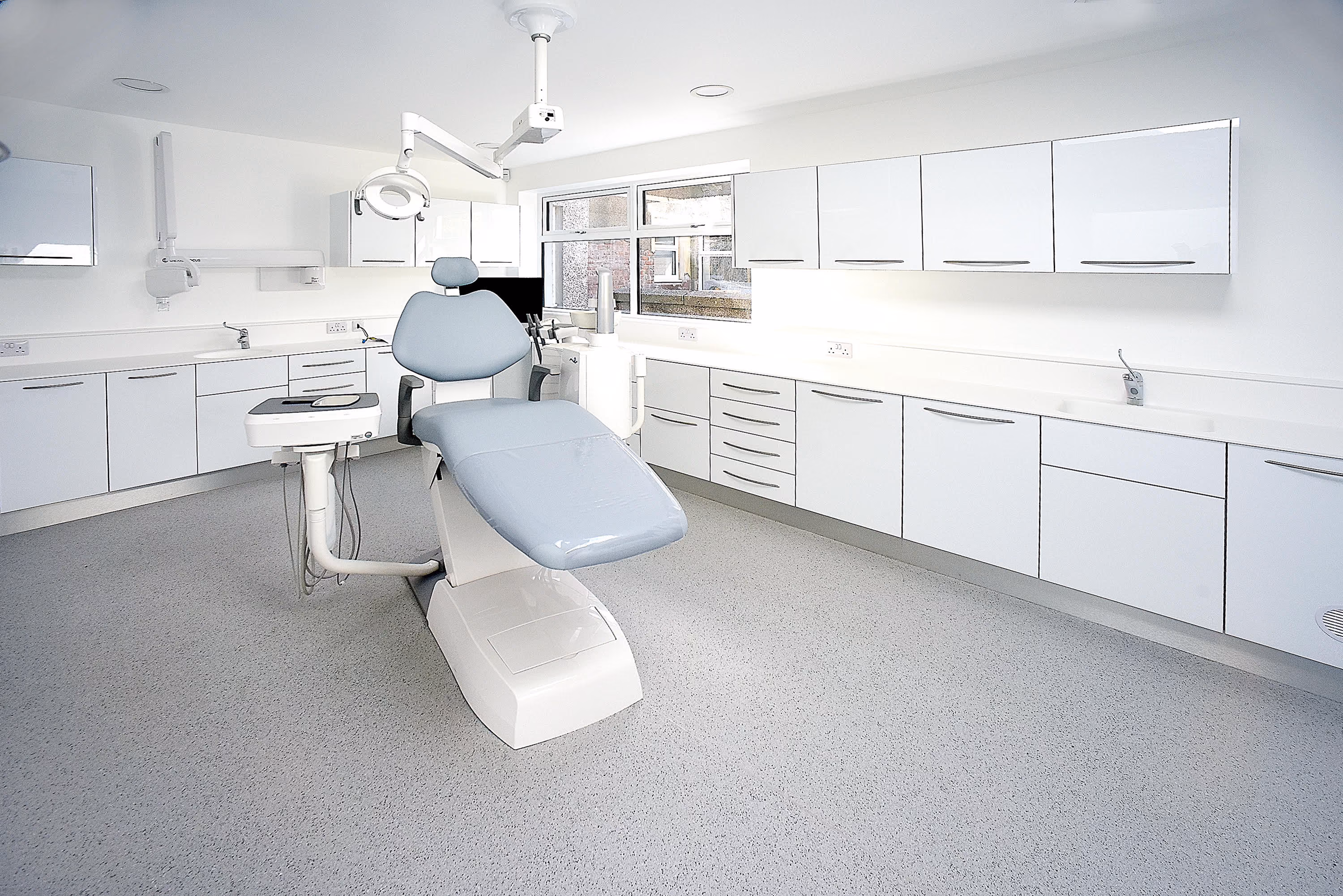 Modern dental clinic room with a blue dental chair, overhead light, and white cabinets.