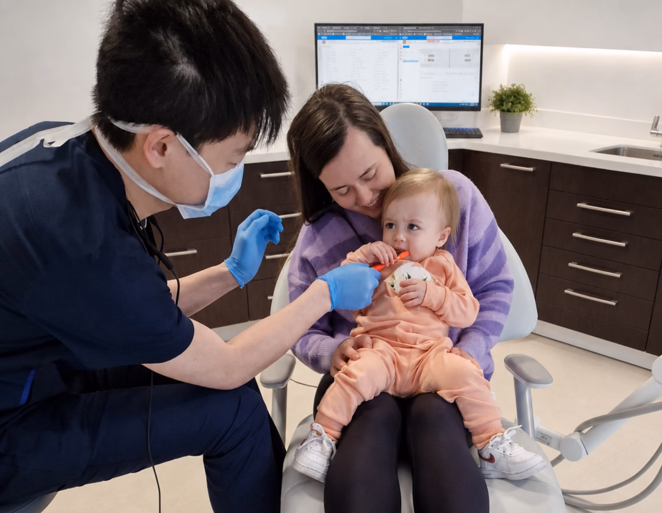 Dentist wearing mask and gloves checking toddler's teeth while child sits on mother's lap in clinic.