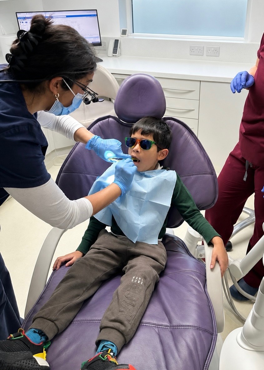 Child wearing sunglasses in dental chair being examined by gloved dentist with dental tool and light.