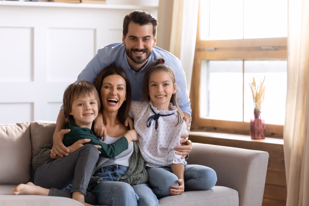 Smiling parents hugging two children on a couch in a bright living room with a window.