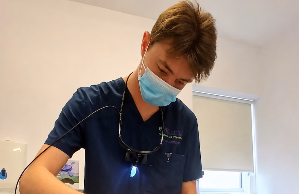 Male dentist in blue scrubs and mask working with dental light on inside a clinic room.