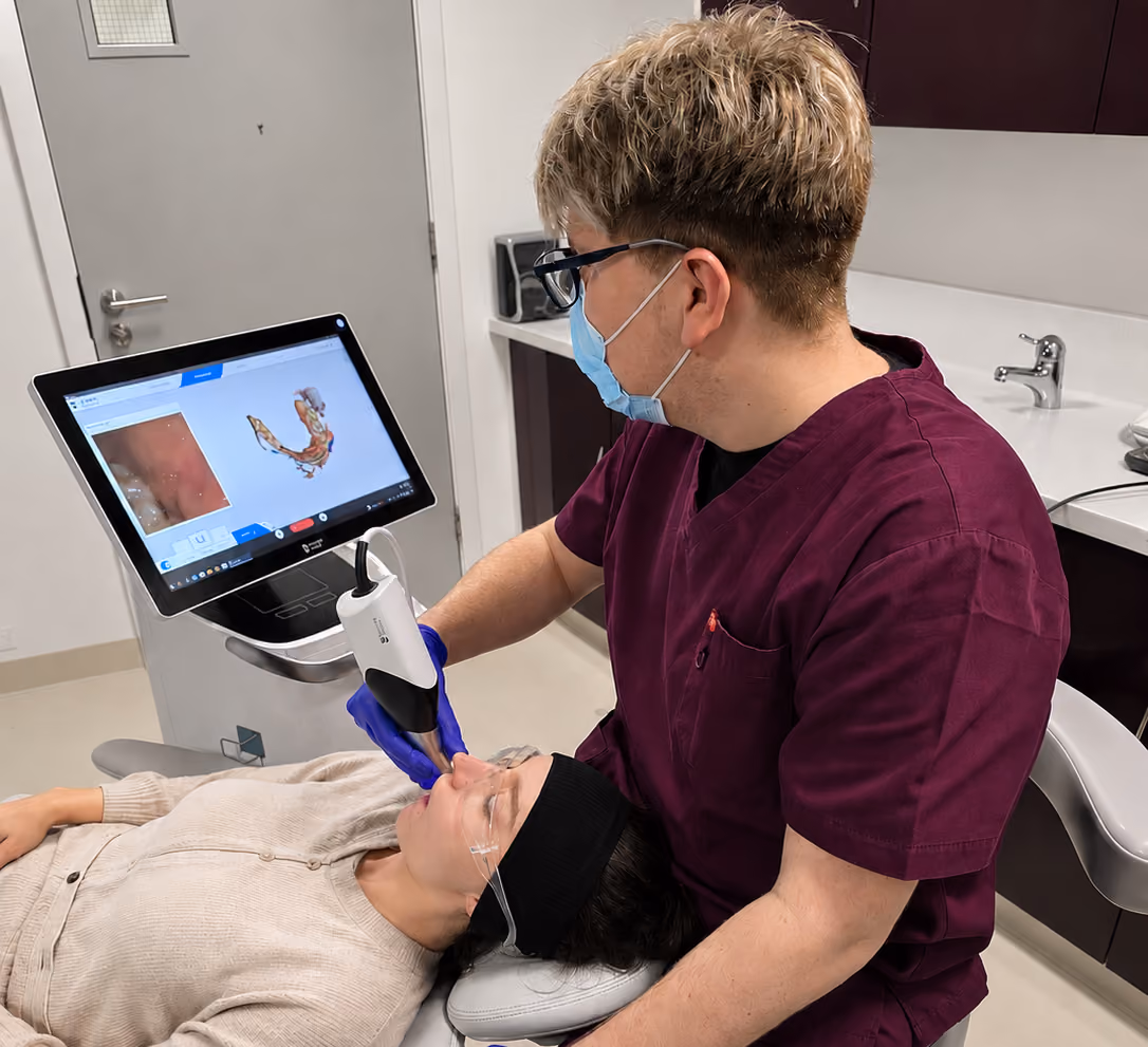 Dentist wearing mask and gloves using digital scanner on patient's teeth, screen shows 3D dental image.