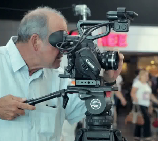 Man looking through the viewfinder of a video camera mounted on a tripod indoors.