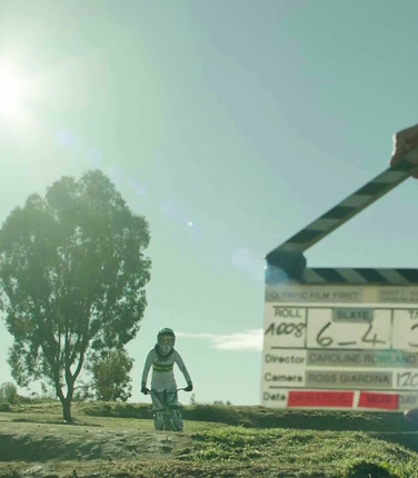 Person riding a bike on a dirt path beside a large tree, with a film clapperboard in the foreground.