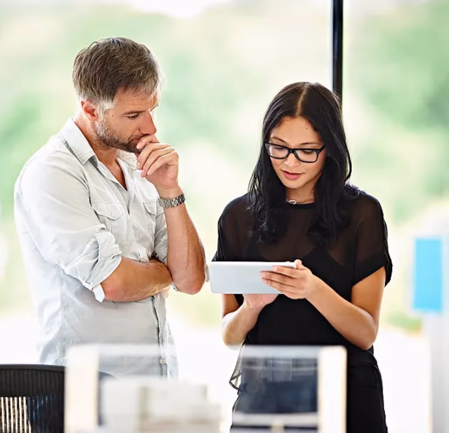 Two colleagues in an office, a woman shows a tablet to a man who is thoughtfully watching.