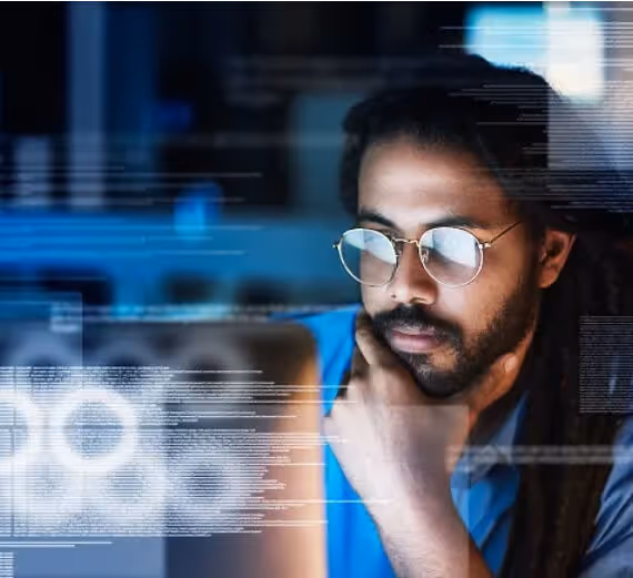 Man with glasses thoughtfully analyzing data or code on a computer screen in a dimly lit room.