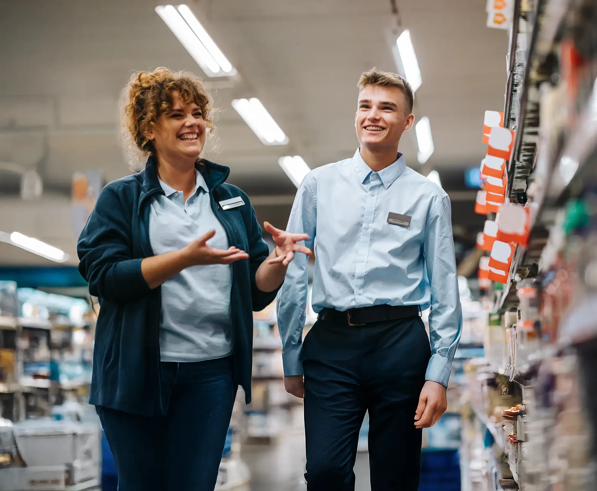 Two smiling store employees walking and talking in a brightly lit supermarket aisle.