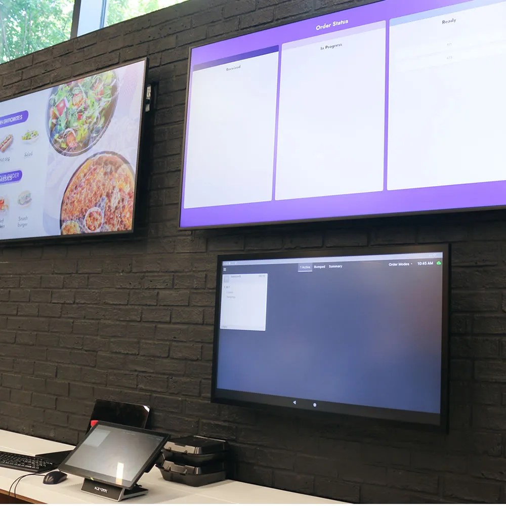 Three screens on a brick wall showing food menu, order status, and order summary in a restaurant setting.