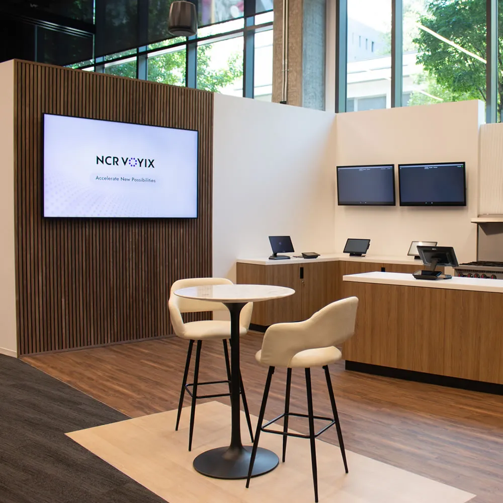 Modern office lounge with tall table, two beige chairs, wood panel wall, and multiple digital screens.