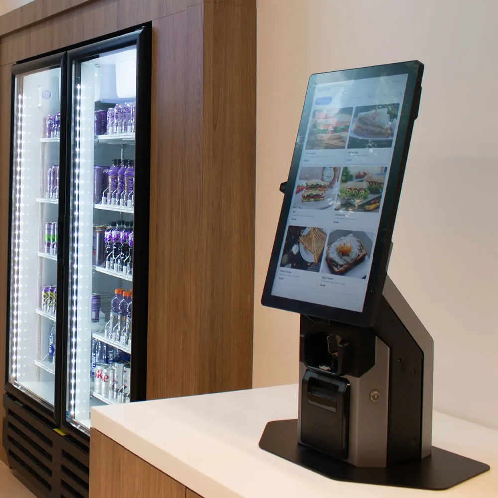 Self-service touchscreen kiosk on counter next to refrigerated beverage cooler with drinks inside.