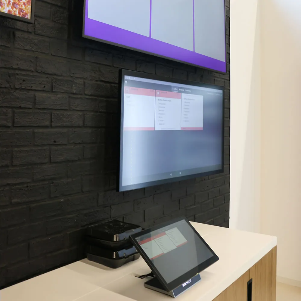 Two black containers and digital order screens on dark brick wall at a white counter in a restaurant.
