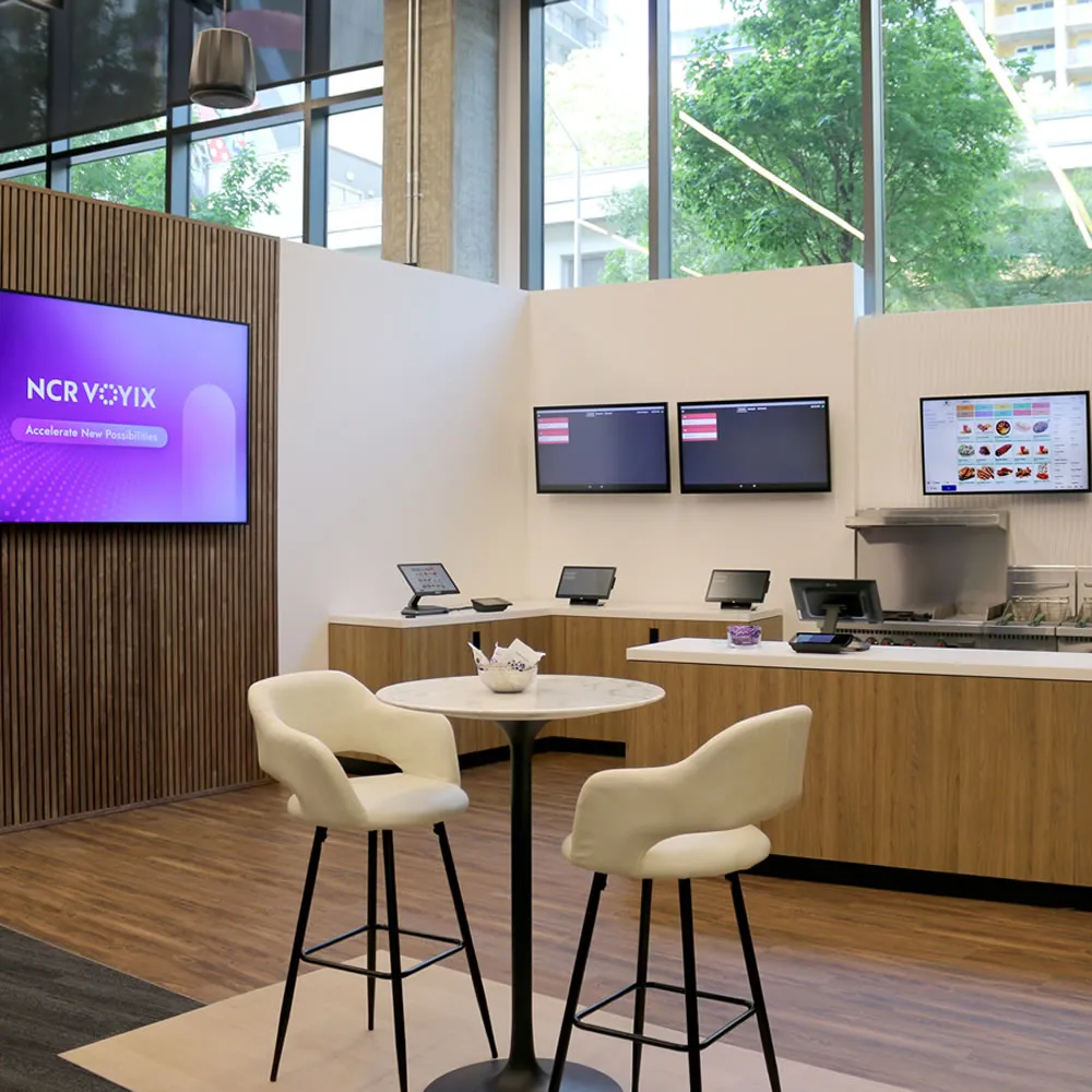 Modern cafe with a round table, two cushioned stools, digital menu screens, and large windows.