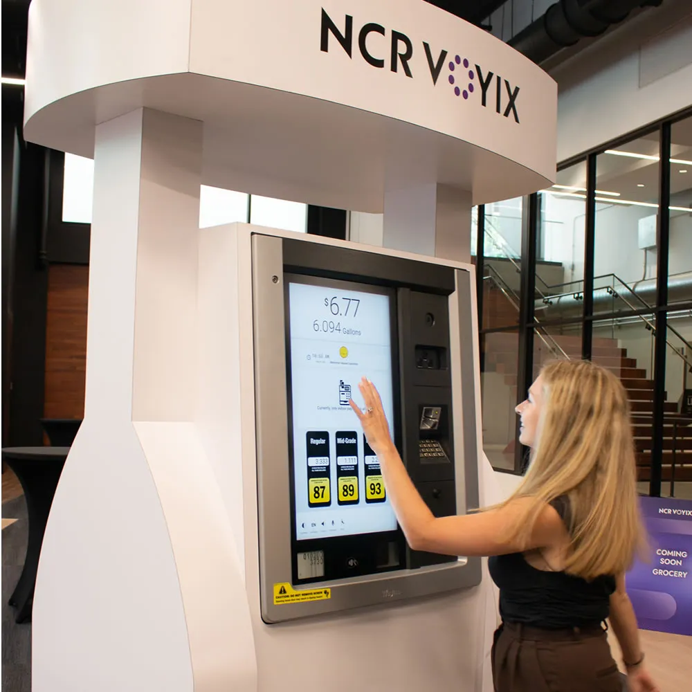 Woman interacting with a large touchscreen display on an NCR Voyix kiosk indoors.
