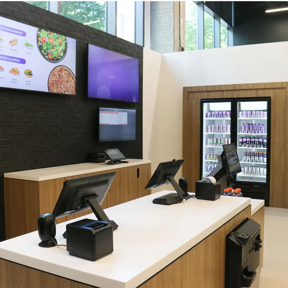 Modern checkout counter with monitors, receipt printers, and beverage coolers stocked with drinks.