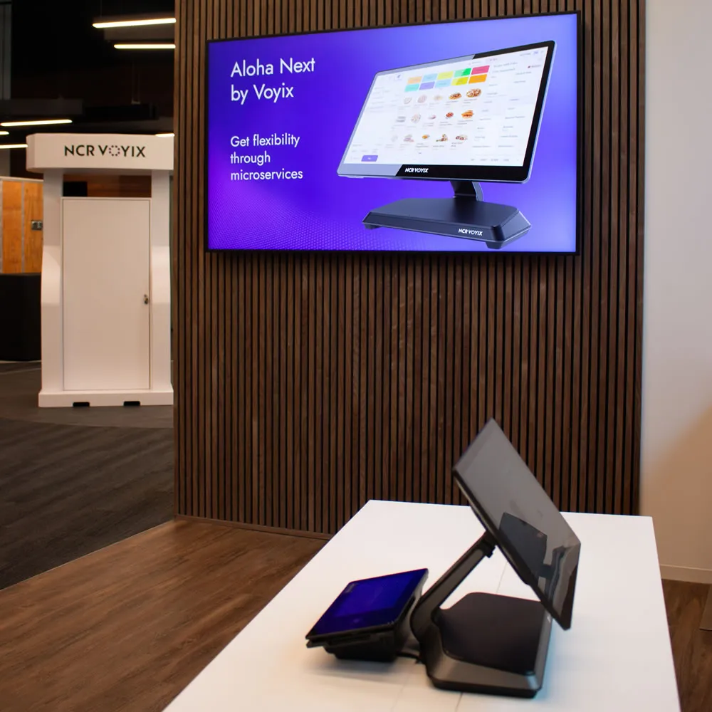 Sales counter with dual touchscreen displays in front of a purple screen showing Aloha Next by Voyix software.
