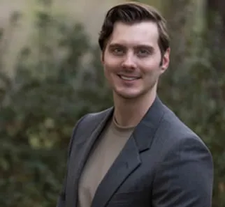 Smiling man with brown hair wearing a gray blazer and beige shirt outdoors with blurred greenery background.
