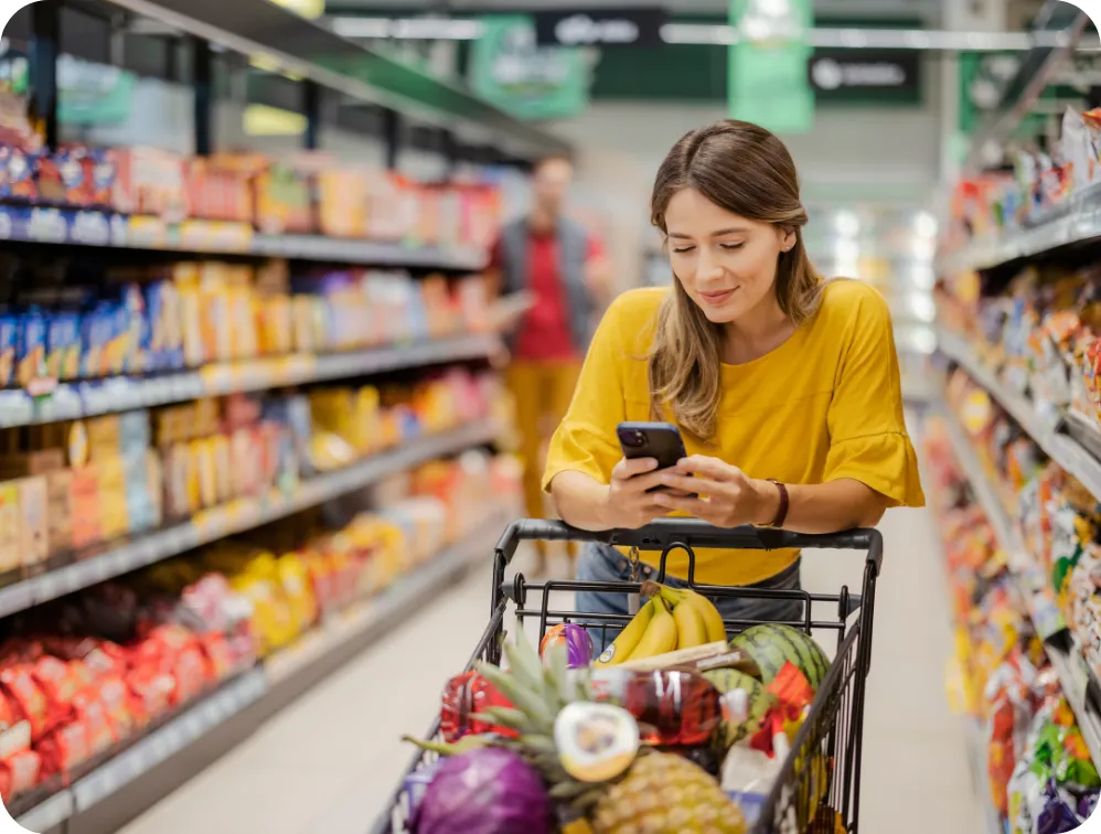 woman consulting a shopping list on her phone standing with a grocery cart filled with produce and other items
