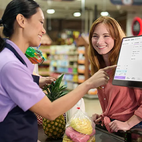 Cashier in purple shirt pointing at a register screen while smiling customer waits with groceries.