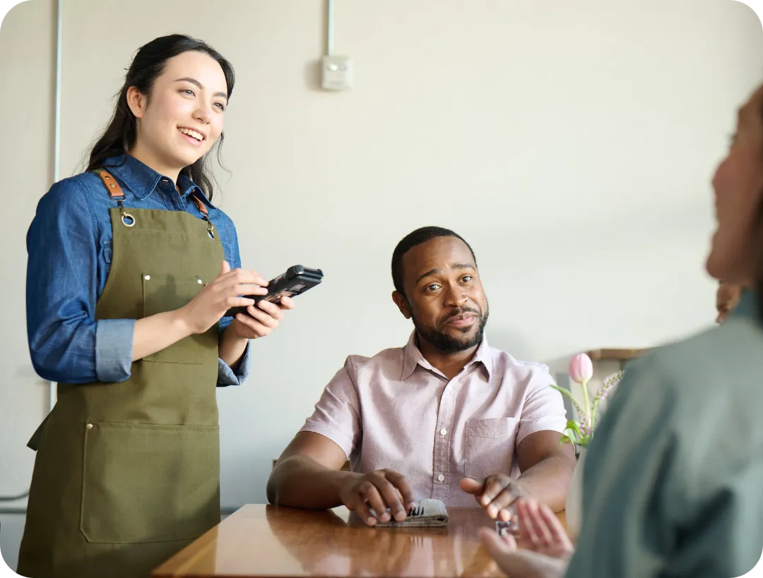 Smiling waitress taking order with a card reader from a seated man at a restaurant table.