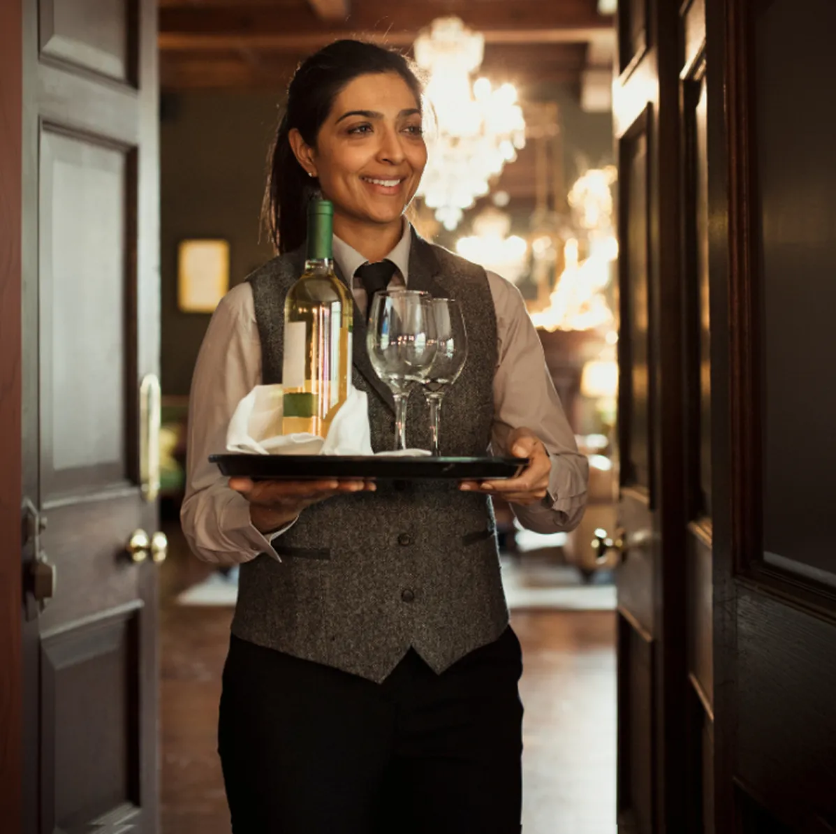 smiling female server in a fine dining restaurant approaching a table with a try of white wine with two glasses