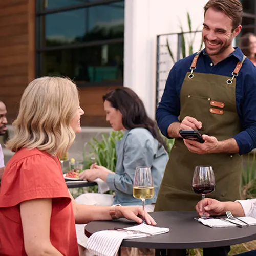 Smiling waiter in an apron taking order from woman seated at outdoor restaurant table with drinks.
