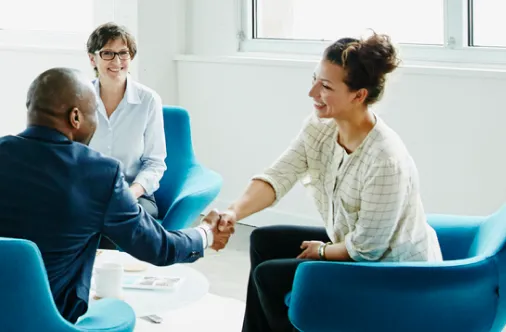 Two people shaking hands in blue chairs while a third person smiles watching the handshake.