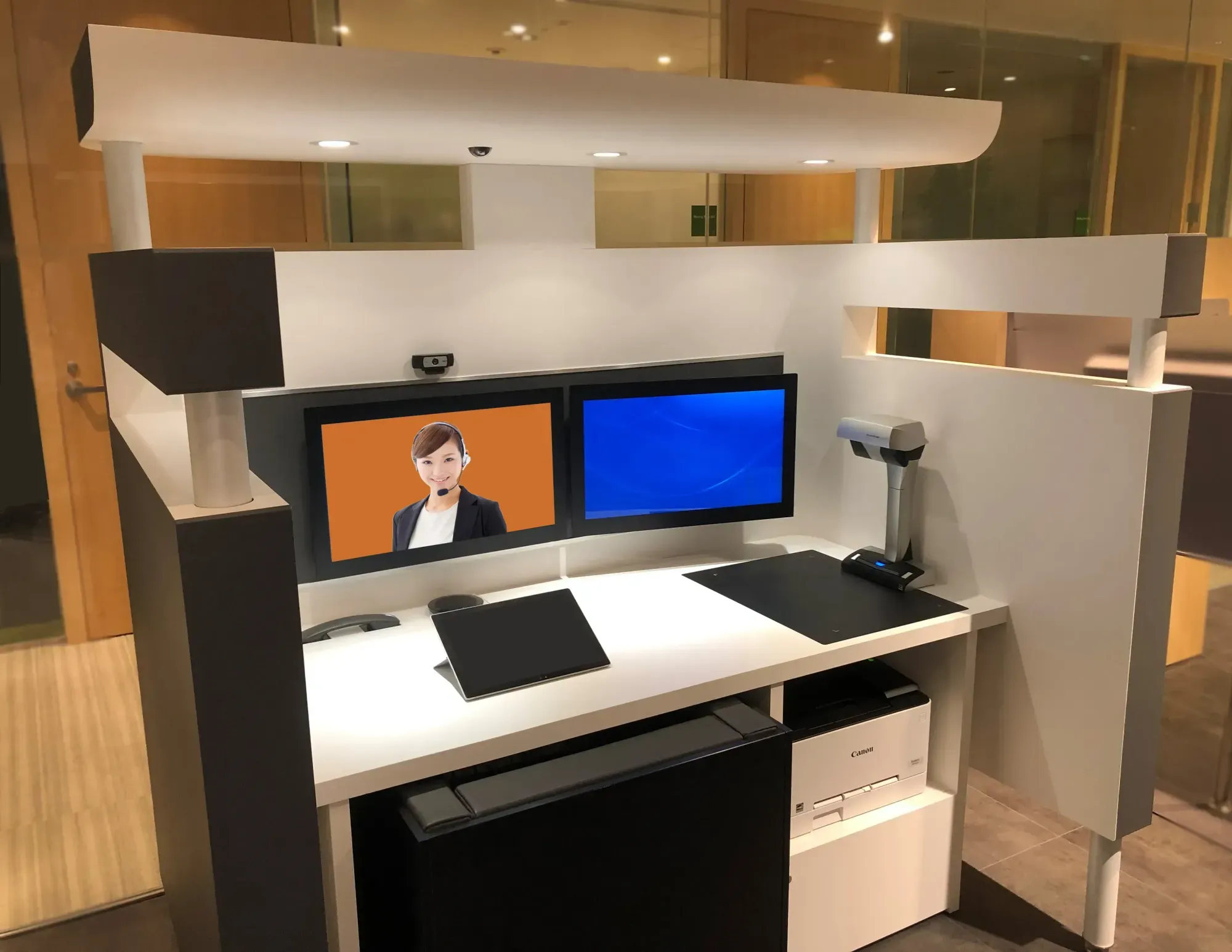 Modern reception desk with dual monitors, one showing a woman with headset, and a Canon printer below.