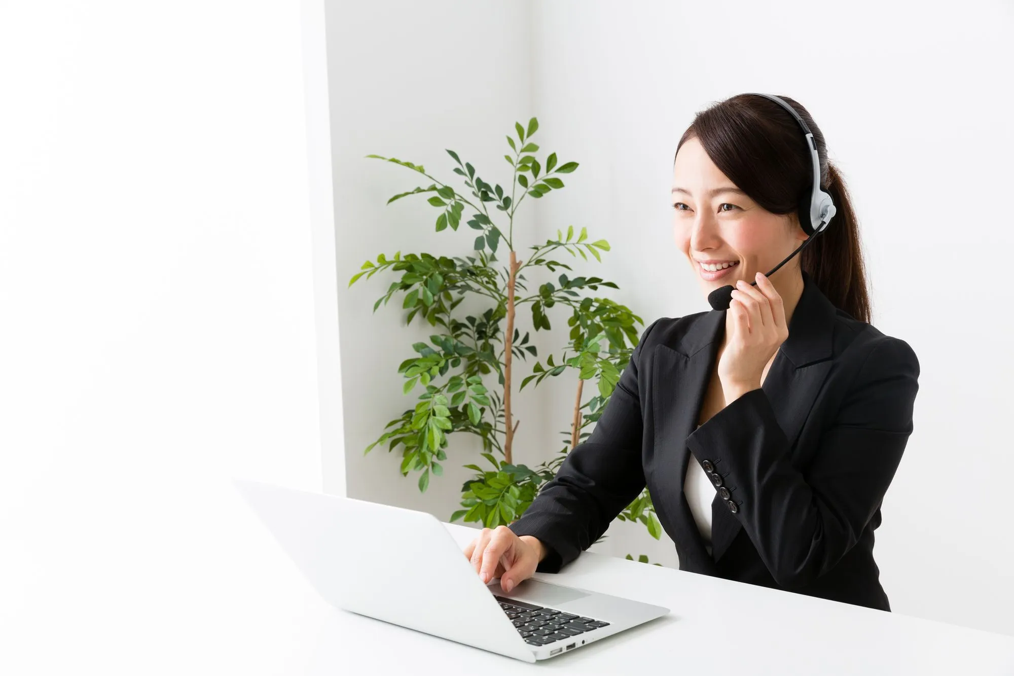 Smiling woman wearing headset working on laptop at white desk with plant in background.