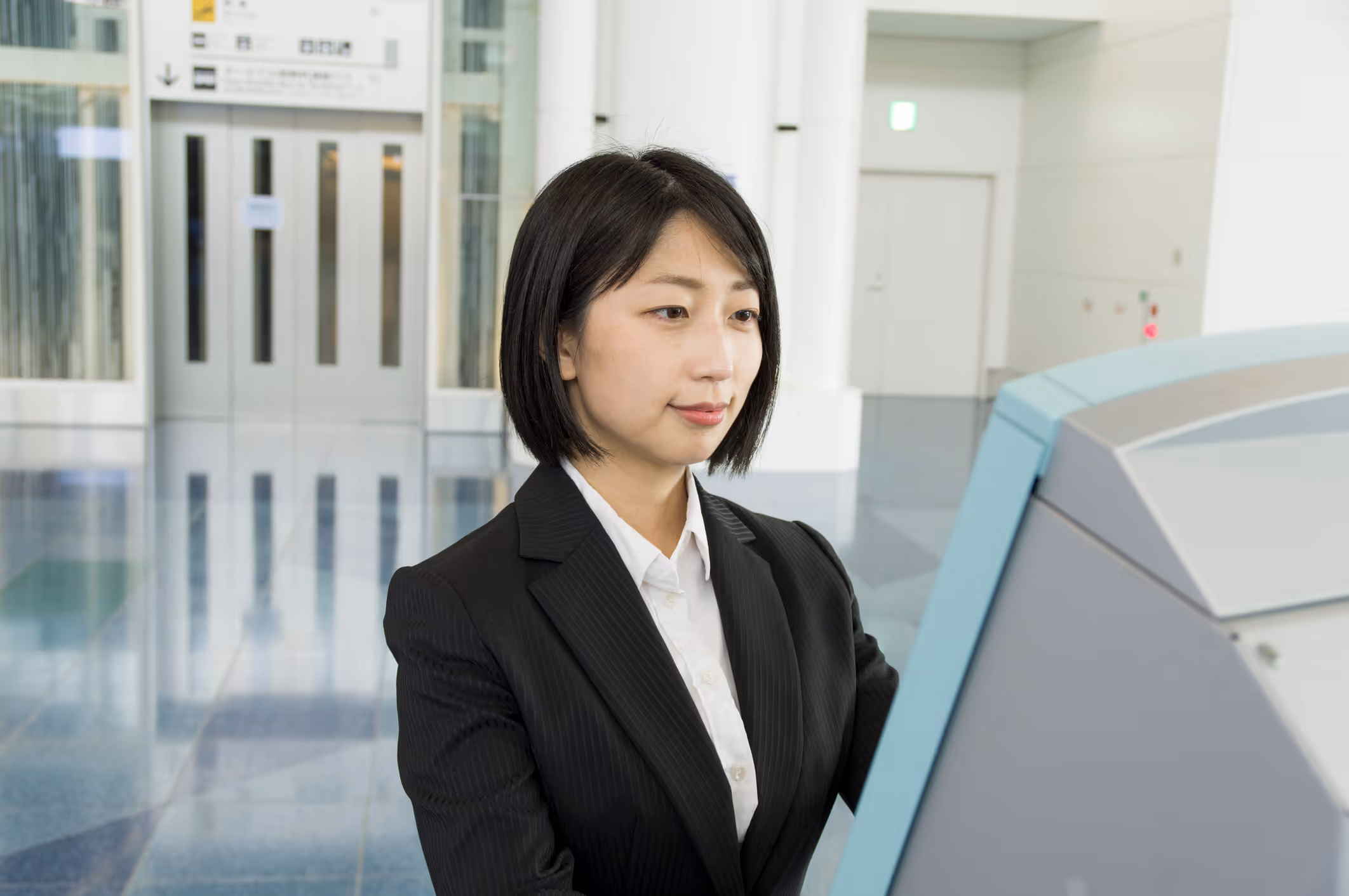 Young woman in black suit using a self-service kiosk in a bright modern building lobby.