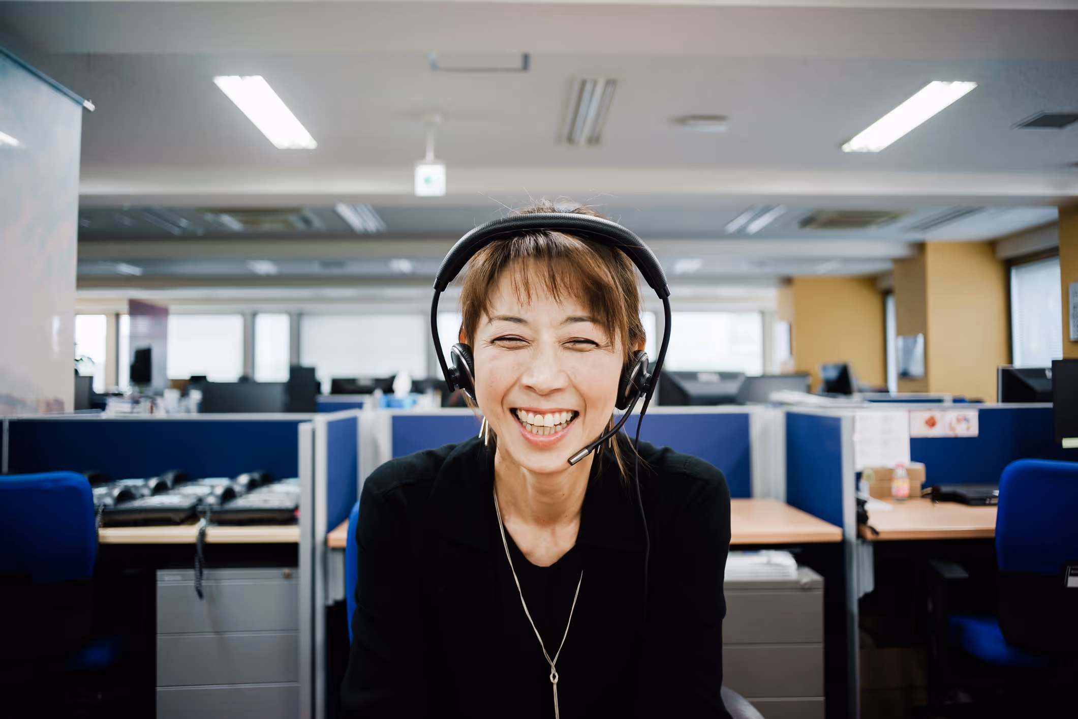 Smiling woman wearing headset sitting in an office cubicle with computers and desks.