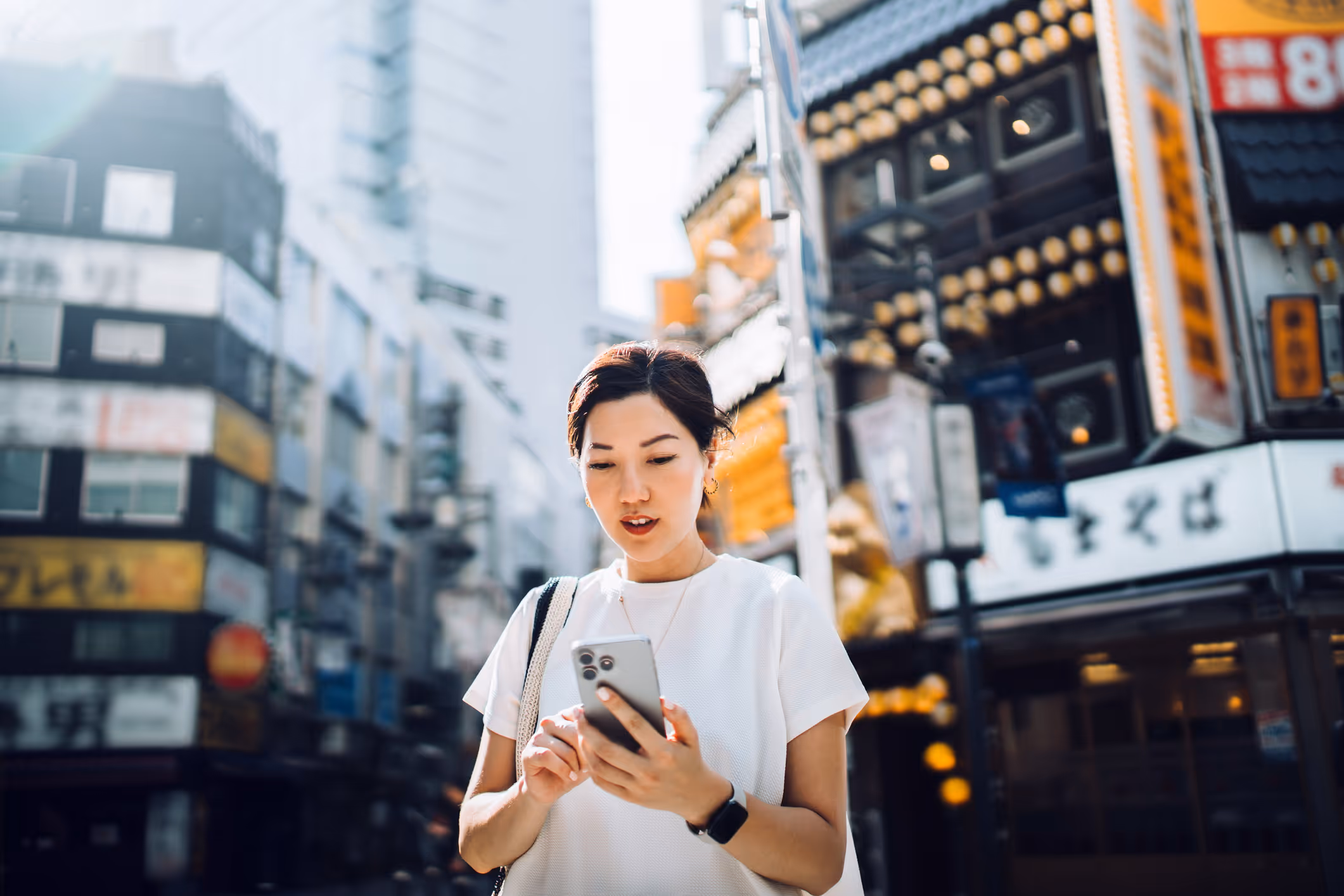 Woman looking at smartphone on a bustling city street with bright signs and buildings.