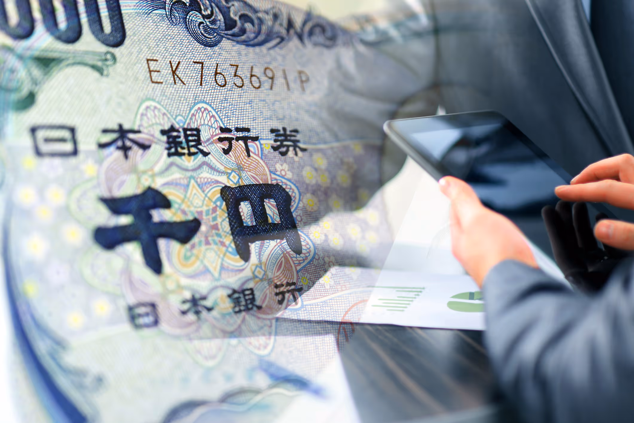 Close-up of Japanese yen banknote overlay and hands using a tablet with financial charts.
