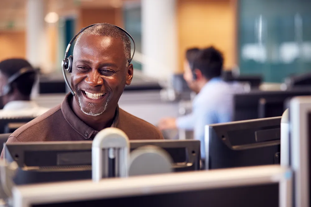 Smiling man wearing headset working at a computer in a busy call center office.