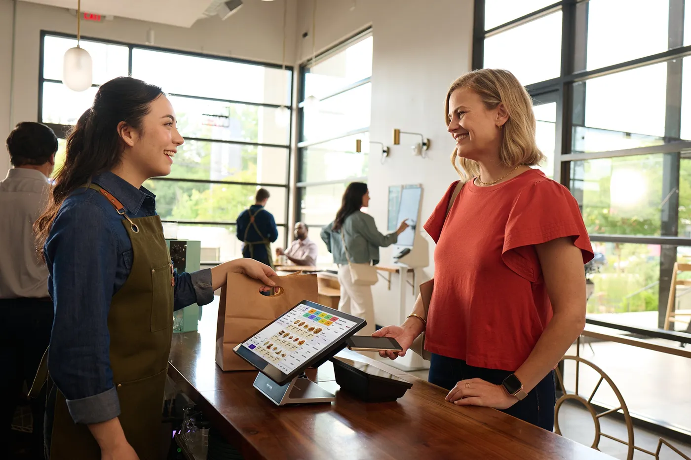 A smiling barista in apron hands a bag to a customer paying with a phone at a café counter.