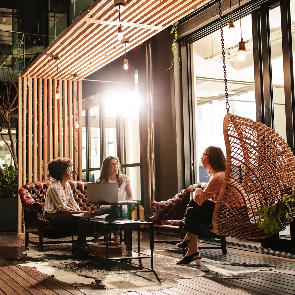 Three women having a casual meeting in a modern room with leather sofas and a hanging wicker chair.