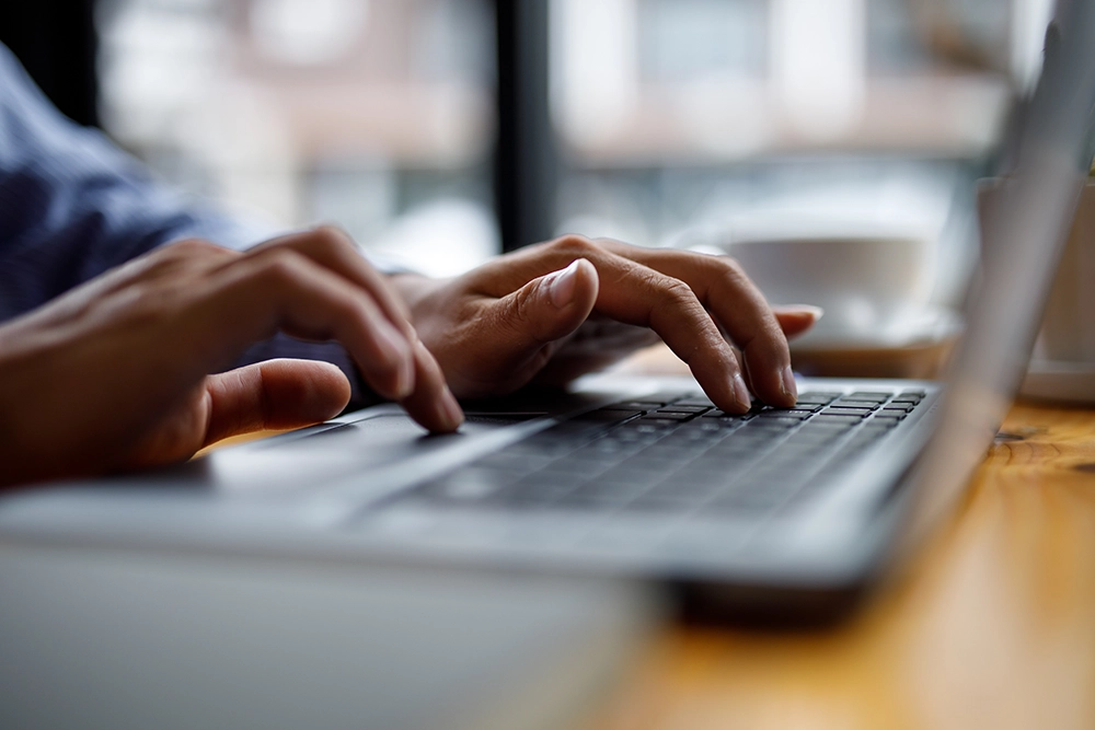 Close-up of hands typing on a laptop keyboard by a window with a blurred coffee cup nearby.