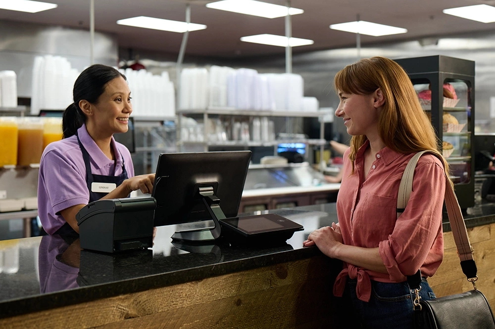 Smiling cashier in a purple shirt helps a female customer at a restaurant counter with a point-of-sale system.