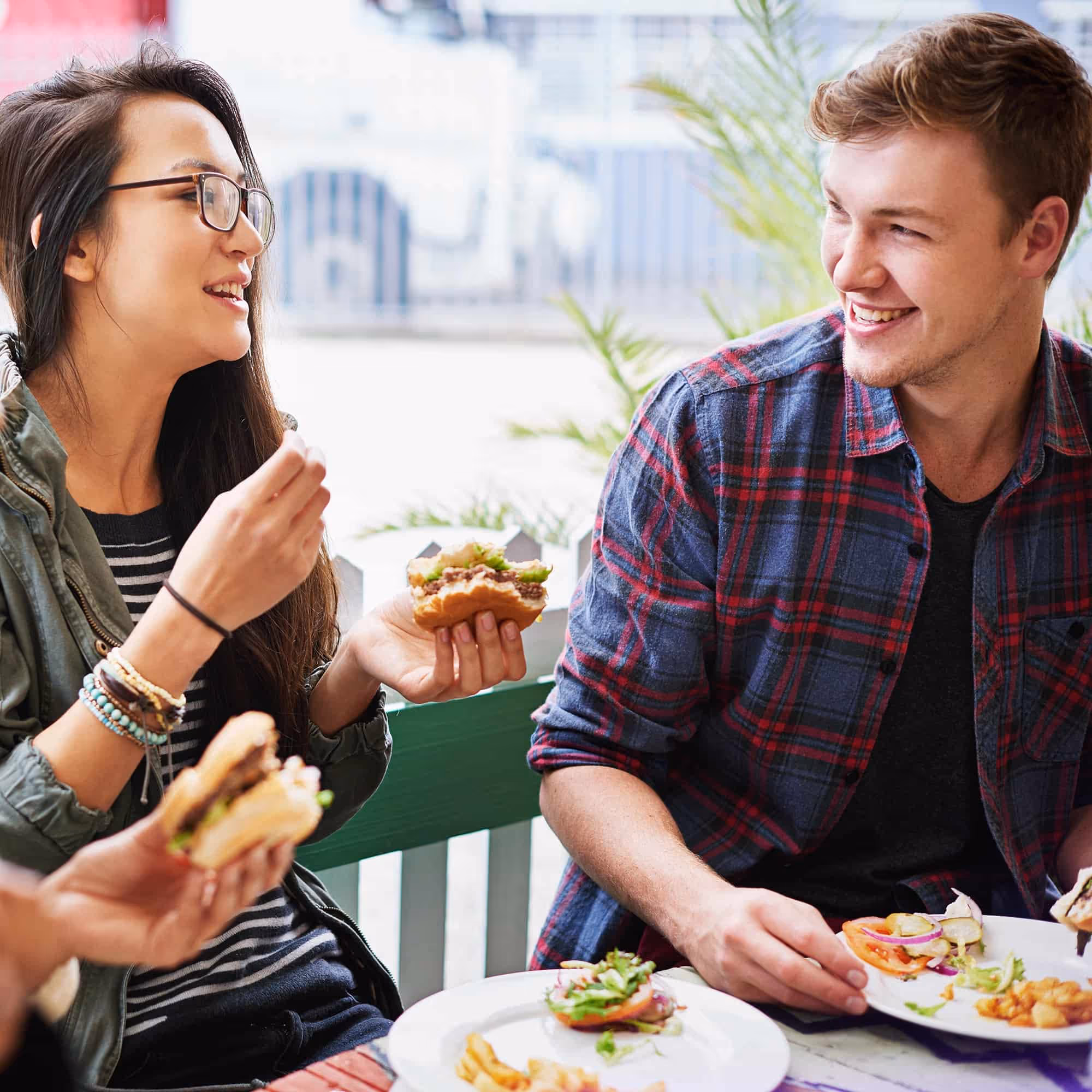A smiling woman with glasses and a man in a plaid shirt enjoying burgers and fries outdoors.