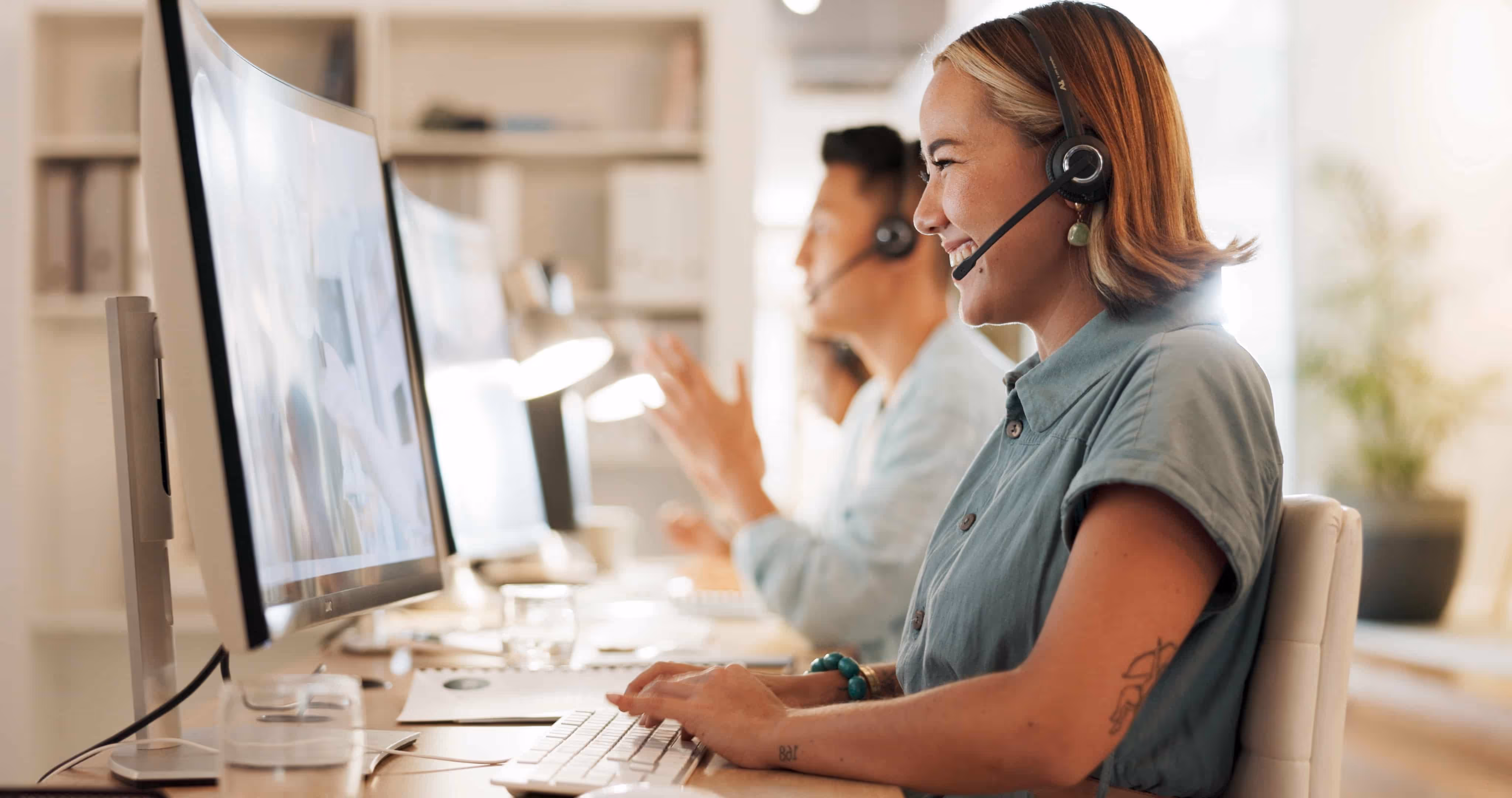 Smiling woman with headset typing at keyboard in a bright office with colleagues using computers.
